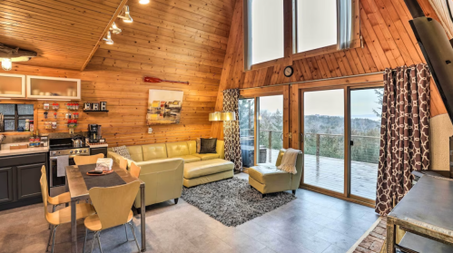 Cozy, modern cabin interior with wooden walls, a large window, and a view of nature. Features a sofa, dining table, and kitchen.